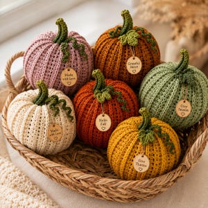 Handmade set of six ribbed crochet pumpkins in dusty pink, burnt orange, sage green, cream, red-orange, and mustard yellow, each with a curved moss green stem, curly vine tendril, and a small wooden charm tag, styled on a fringed canvas runner with autumn leaves and a lit candle.