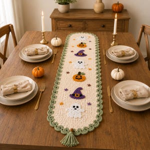 May include: A cream-colored crocheted table runner with a scalloped sage green border and a tassel at the end. The runner features appliqued motifs of white ghosts, orange jack-o'-lanterns, purple witch hats, and small colorful stars.