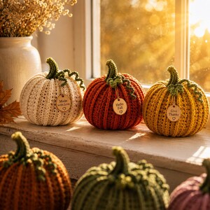 Handmade set of six ribbed crochet pumpkins in dusty pink, burnt orange, sage green, cream, red-orange, and mustard yellow, each with a curved moss green stem, curly vine tendril, and a small wooden charm tag, styled on a fringed canvas runner with autumn leaves and a lit candle.