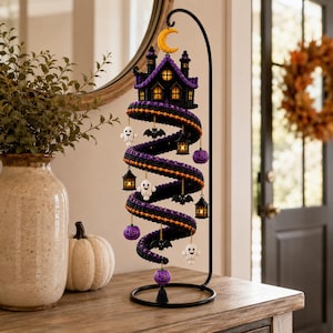May include: A decorative Halloween spiral display hanging from a black metal stand. The structure features a purple and black crocheted spiral adorned with small white ghosts, black bats, purple pumpkins, and miniature lanterns with warm yellow lights.