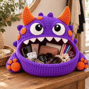 Puede incluir: Una cesta de almacenamiento de ganchillo con forma de monstruo morado, con cuernos y manchas naranjas, dos grandes ojos blancos con pupilas negras y una boca ancha llena de dientes triangulares blancos. El interior contiene varios objetos.