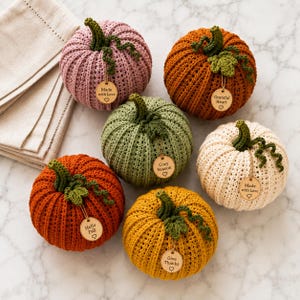 Handmade set of six ribbed crochet pumpkins in dusty pink, burnt orange, sage green, cream, red-orange, and mustard yellow, each with a curved moss green stem, curly vine tendril, and a small wooden charm tag, styled on a fringed canvas runner with autumn leaves and a lit candle.