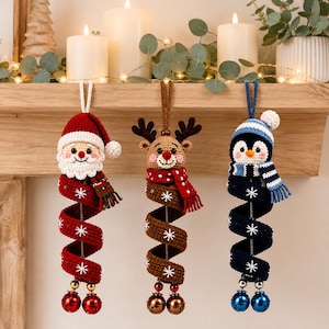 Könnte beinhalten: Drei gehäkelte Hängedekorationen in Form eines Weihnachtsmanns, eines Rentiers und eines Pinguins. Jede Figur hat einen spiralförmigen Körper mit weißen Schneeflockenstickereien und zwei Glöckchen am unteren Ende. Sie hängen an farbigen Schlaufen.