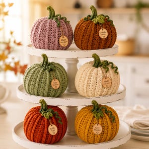 Handmade set of six ribbed crochet pumpkins in dusty pink, burnt orange, sage green, cream, red-orange, and mustard yellow, each with a curved moss green stem, curly vine tendril, and a small wooden charm tag, styled on a fringed canvas runner with autumn leaves and a lit candle.