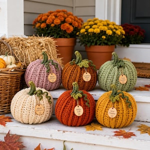 Handmade set of six ribbed crochet pumpkins in dusty pink, burnt orange, sage green, cream, red-orange, and mustard yellow, each with a curved moss green stem, curly vine tendril, and a small wooden charm tag, styled on a fringed canvas runner with autumn leaves and a lit candle.