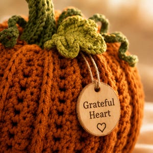 Handmade set of six ribbed crochet pumpkins in dusty pink, burnt orange, sage green, cream, red-orange, and mustard yellow, each with a curved moss green stem, curly vine tendril, and a small wooden charm tag, styled on a fringed canvas runner with autumn leaves and a lit candle.