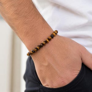 May include: A beaded bracelet featuring smooth, round tiger's eye gemstones. The beads display a natural chatoyant luster with alternating bands of golden-brown and dark brown colors, strung together in a single continuous circular strand.