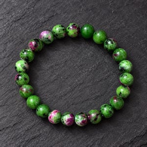 May include: A circular beaded bracelet composed of polished, spherical stones. The beads feature a mottled pattern of vibrant green and deep magenta-pink, with small black inclusions distributed throughout the surface of each individual stone.