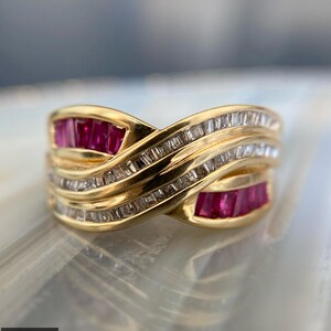 May include: A gold ring featuring a crossover design with rows of channel-set baguette-cut rubies and clear baguette-cut diamonds. The band has a polished finish, with the red rubies and clear diamonds arranged in a flowing, intertwined pattern.