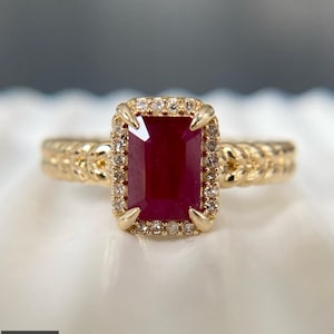 May include: A gold ring featuring a rectangular, deep red ruby center stone surrounded by a halo of small, clear, round-cut diamonds. The ring band has a textured, braided metal design, and the center stone is secured by four gold prongs.