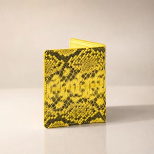 May include: A bi-fold wallet featuring a vibrant yellow and black snakeskin pattern. The exterior displays a textured, scale-like appearance with a repeating dark organic print over a bright yellow base, finished with visible stitching along the edges.