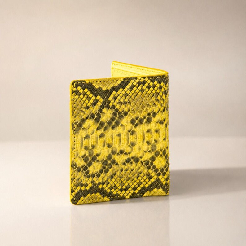 May include: A bi-fold wallet featuring a vibrant yellow and black snakeskin pattern. The exterior displays a textured, scale-like appearance with a repeating dark organic print over a bright yellow base, finished with visible stitching along the edges.