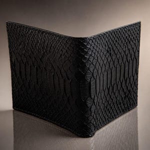 May include: A bi-fold wallet featuring a black exterior with a textured snakeskin pattern. The wallet is positioned upright in an open orientation, showcasing the detailed scale-like surface and the clean, straight edges of the folded construction.