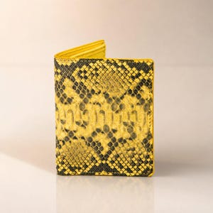 May include: A bi-fold wallet featuring a vibrant yellow and black snakeskin pattern. The exterior displays a detailed scale texture across the surface, while the interior lining is a solid, bright yellow color visible at the top fold.