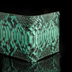 Puede incluir: Una cartera plegable con un patrón de piel de serpiente texturizada en tonos verde azulado vibrante y negro. El material muestra una superficie distintiva similar a escamas con un diseño orgánico repetitivo y un contraste marcado.
