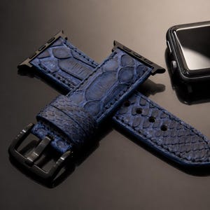 May include: A deep blue watch strap with a textured, scale-like pattern and a black metal buckle, positioned next to a black smartwatch case. The strap features a matte finish and includes black metal adapters for attachment to the watch device.