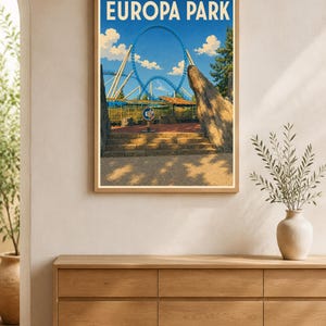Może przedstawiać: Oprawiony wydruk artystyczny przedstawiający kolorową ilustrację kolejki górskiej w Europa Park z dużą niebieską pętlą, kamiennymi filarami i błękitnym niebem. Napis EUROPA PARK znajduje się w górnej części grafiki białymi literami.