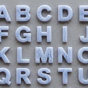 May include: A collection of twenty uppercase letters from A to U, crafted from light blue and white banded agate stone. Each letter features a smooth, polished surface with natural, wavy, and layered mineral patterns in shades of pale blue and white.
