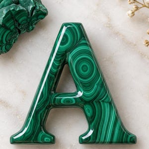 May include: A capital letter A shaped object featuring a polished surface with a deep green and black banded malachite pattern. The letter has a smooth, glossy finish with distinct concentric circular and wavy lines characteristic of the stone.