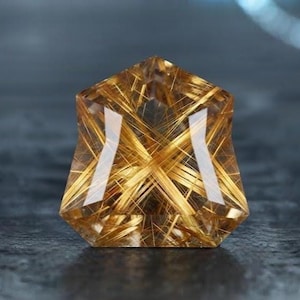 May include: A faceted shield-shaped rutilated quartz gemstone featuring a clear, golden-brown body filled with numerous intersecting, needle-like golden rutile inclusions that create a distinct starburst pattern throughout the transparent stone.
