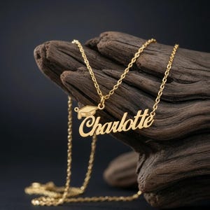 Könnte beinhalten: Eine goldfarbene Metallhalskette mit einem Namensschild in Schreibschrift, auf dem Charlotte steht. An dem Namensschild ist ein kleiner Anhänger in Form einer Doktorhut-Kappe mit der eingravierten Jahreszahl 2024 befestigt.