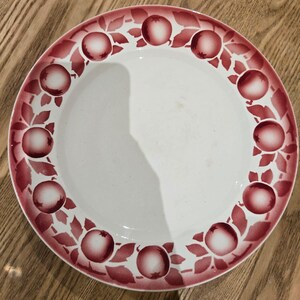 Peut inclure: Une assiette ronde en céramique blanche avec une bordure décorative composée de grenades et de feuilles rouge foncé. Le centre de l'assiette est blanc uni, tandis que le bord est orné d'un motif répétitif de fruits et de feuillage rouge.