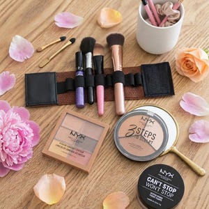 Puede incluir: Una colección de productos de maquillaje que incluye una paleta NYX 3 Steps to Sculpt, un envase de polvos Can't Stop Won't Stop, una paleta de contorno cuadrada, un organizador de brochas marrón con cuatro brochas y un vaso blanco con tres brochas adicionales.