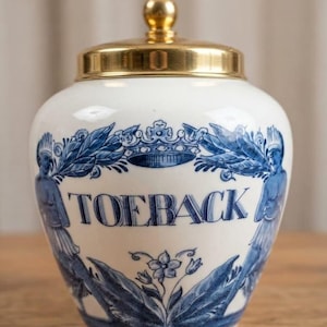 Peut inclure: Un pot en céramique blanche avec un motif décoratif bleu comprenant le texte TOEBACK, une couronne centrale et deux personnages sur les côtés. Le pot est surmonté d'un couvercle en métal doré poli doté d'une petite poignée sphérique.