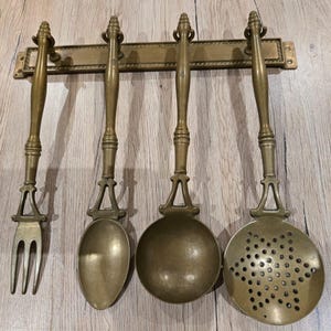 Peut inclure: Un ensemble de quatre ustensiles de cuisine en laiton suspendus à une barre murale assortie. Le set comprend une fourchette à deux dents, une cuillère pleine, une louche profonde et une écumoire perforée, dotées de longs manches tournés.