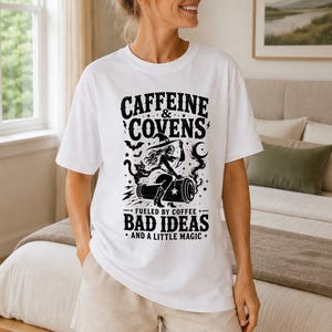 May include: A white short-sleeve t-shirt featuring a black graphic print. The text reads CAFFEINE & COVENS, FUELED BY COFFEE, BAD IDEAS, AND A LITTLE MAGIC. The illustration depicts a witch riding a coffee mug with a cat, bats, and stars.