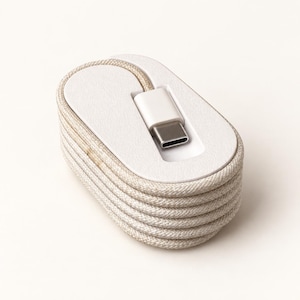 Op de afbeelding: Een opgerolde beige gevlochten oplaadkabel gewikkeld rond een witte ovale plastic spoel. De kabel is voorzien van een witte USB-C-connector die stevig in een verzonken gleuf op het bovenoppervlak van de spoel zit voor een georganiseerde opslag.