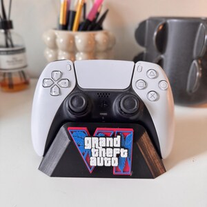 Può includere: Un controller di gioco wireless bianco e nero appoggiato su un supporto in plastica nera. Il supporto presenta un logo Grand Theft Auto V in rosso, blu e bianco, con il testo Grand Theft Auto chiaramente visibile sulla parte anteriore.