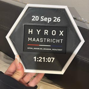 Op de afbeelding: Een zeshoekige plaquette met een witte rand en een zwart midden met de tekst 20 Sep 26, HYROX MAASTRICHT, een rode, witte en blauwe lijn, de tekst OPEN_DOUBLES_SEASON_2026/2027 en de tijd 1:21:07 in witte letters.