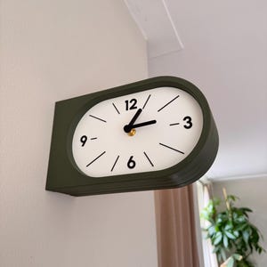 Op de afbeelding: Een wandklok met een donker olijfgroen, afgerond rechthoekig frame en een witte wijzerplaat. De klok heeft zwarte uur- en minuutwijzers, met zwarte cijfers op de posities 12, 3, 6 en 9, en eenvoudige zwarte lijnen voor de overige uren.