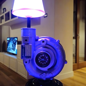 Op de afbeelding: Een tafellamp gemaakt van een zilverkleurige metalen turboladerbehuizing. De lamp is voorzien van een gloeiende roze lampenkap aan de bovenkant, een analoge klok in het midden en een zwarte ronde poelie als voet aan de onderkant.