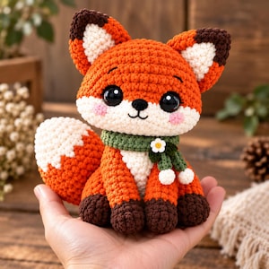 May include: A small crocheted fox plush toy with bright orange and white yarn, featuring large black eyes, pink cheeks, and dark brown paws. The fox wears a green crocheted scarf adorned with a small white flower and two white pom-poms.