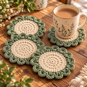 May include: A set of three round, crocheted coasters featuring a cream-coloured centre and a scalloped sage green border. One coaster supports a speckled ceramic mug filled with a light brown beverage, while two others lie flat nearby.