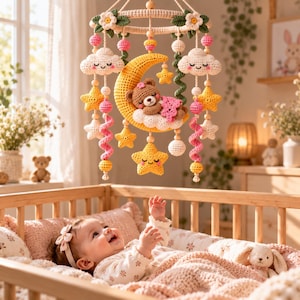 May include: A crochet baby mobile featuring a central yellow crescent moon with a sleeping brown bear, surrounded by hanging white clouds, yellow stars, and pink spiral elements, all suspended from a circular frame decorated with white flowers.