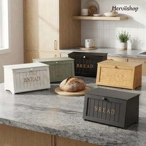 May include: Four rectangular bread boxes in white, sage green, black, and natural wood tones, each featuring a hinged lid with a small knob and the word BREAD embossed on the front. A round loaf of bread sits on a wooden board in the center.