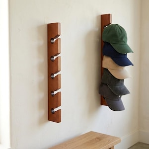 May include: Two vertical wooden wall-mounted racks with five metal hooks each. One rack is empty, while the other holds a stack of six baseball caps in various colors including green, navy blue, tan, dark gray, and light gray.