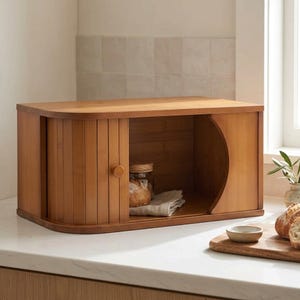 May include: A rectangular bamboo bread box with a curved roll-top door featuring a small wooden knob. The interior contains a glass jar and a folded cloth napkin. The exterior has a light brown wood grain finish with rounded side edges.
