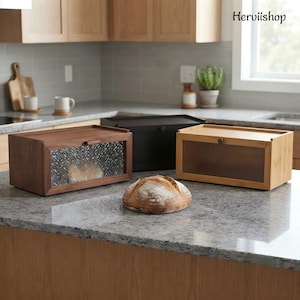 May include: Three rectangular bread boxes in dark wood, black, and light wood finishes. The dark wood box has a textured glass front, the black box is solid, and the light wood box features a ribbed glass front. A round loaf of bread sits in front.
