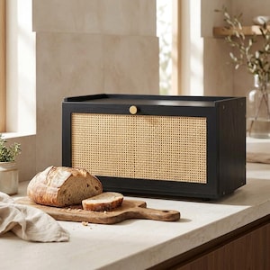 May include: A rectangular black bread box with a natural woven cane front panel and a small circular gold knob. A loaf of sourdough bread sits on a wooden cutting board in front of the container, with a slice resting beside it.