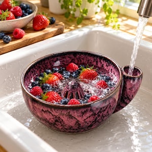 Könnte beinhalten: Ein rundes, tiefes Beeren-Sieb aus Keramik mit einer gesprenkelten magenta-schwarzen Glasur, ausgestattet mit einem zentralen Abflusskegel und einem seitlichen Griff. Die Schale ist mit frischen roten Erdbeeren und Blaubeeren gefüllt.