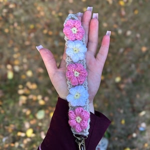 May include: A crochet dummy clip featuring a beige strap decorated with five alternating pink and white flowers, each with a small yellow centre. The strap is finished with a silver-toned metal clip at the bottom for secure attachment.