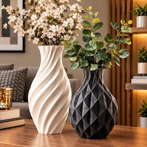 May include: A tall white vase with a spiraling vertical texture containing white flowers and a shorter black vase with a geometric faceted surface containing green leafy branches. Both vases are positioned side by side on a wooden surface.