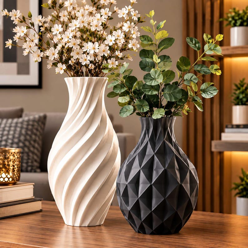 May include: A tall white vase with a spiraling vertical texture containing white flowers and a shorter black vase with a geometric faceted surface containing green leafy branches. Both vases are positioned side by side on a wooden surface.
