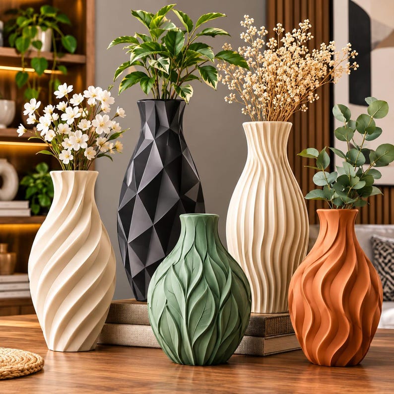 May include: A collection of five decorative vases in various shapes and colors, including a white spiral vase, a black geometric vase, a green leaf-textured vase, a tall cream ribbed vase, and a terracotta-colored wavy vase, all holding plants.