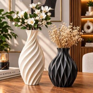 May include: A tall white vase with a spiraling vertical texture containing white magnolia flowers and a shorter black vase with a geometric diamond-patterned surface containing dried beige filler flowers, both standing on a wooden surface.