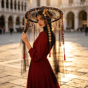 May include: A deep red long-sleeved dress paired with a black head covering and a wide-brimmed black hat. The hat features hanging gold-toned circular coins, thin gold chains, and long black sheer veils accented with red beaded tassels.